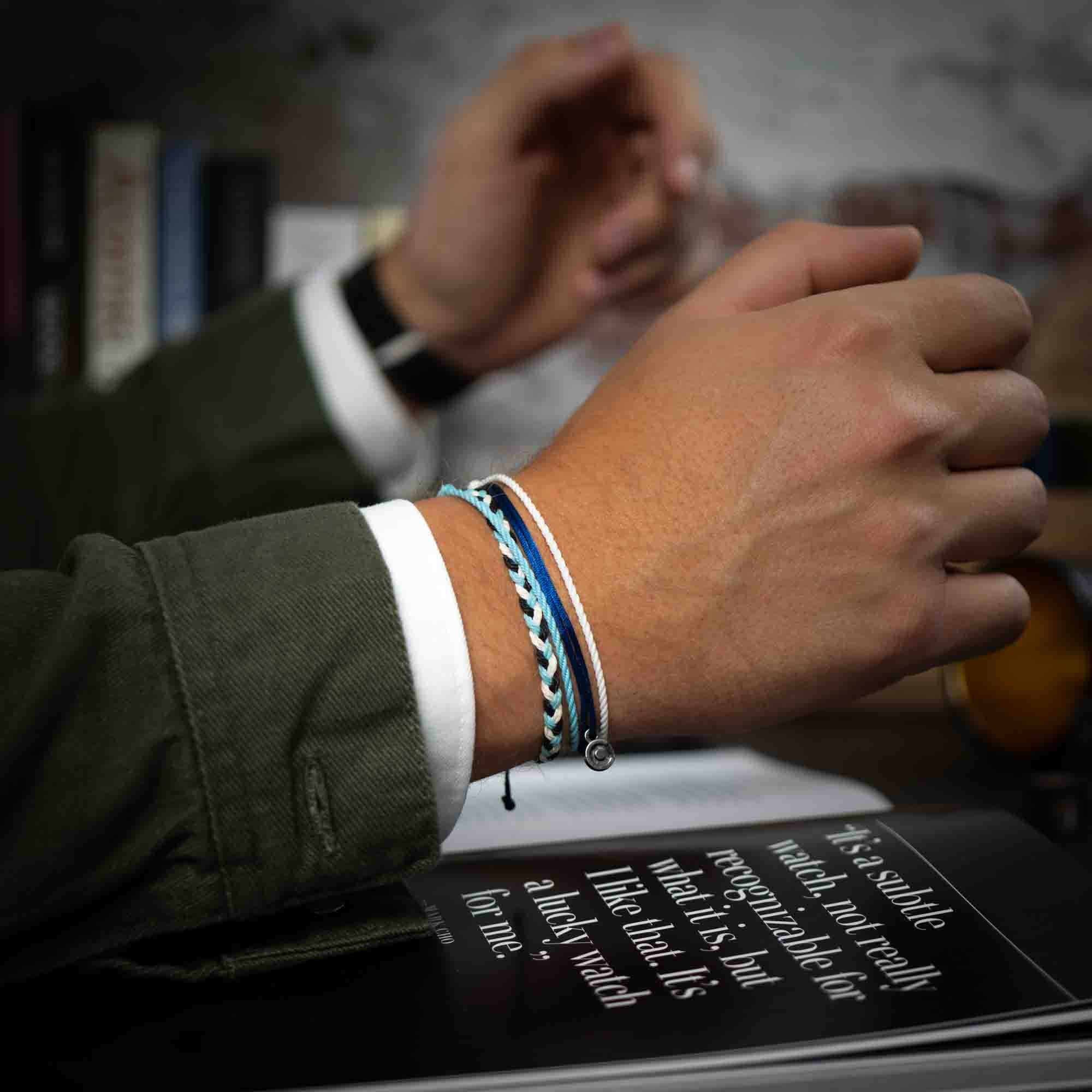A person's wrist wearing a white and blue wax-coated polyester bracelet with a Chibuntu charm, displayed in front of a book.