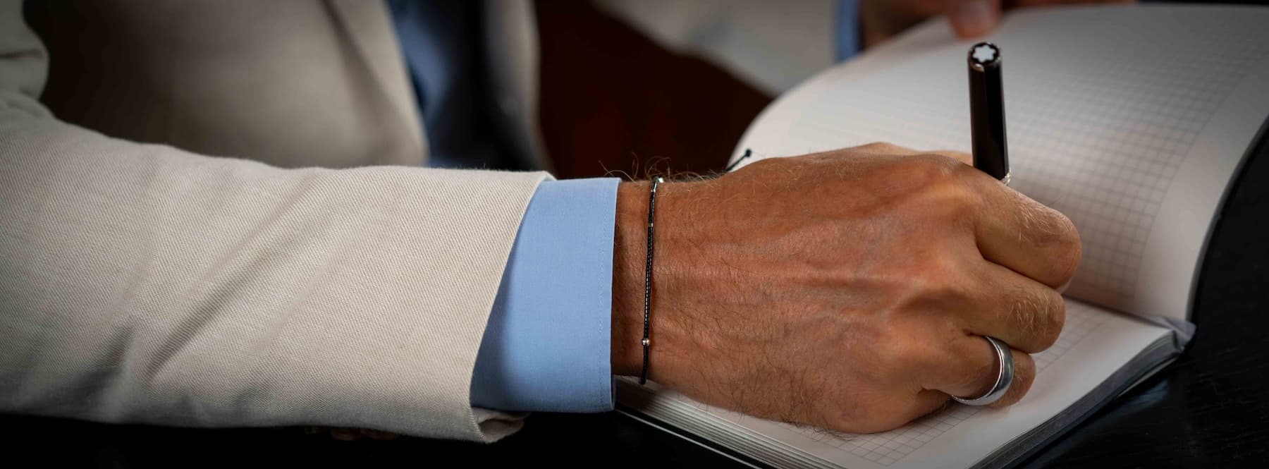 Hand writing in a notebook with a Montblanc pen, wearing a light blue shirt and beige jacket.