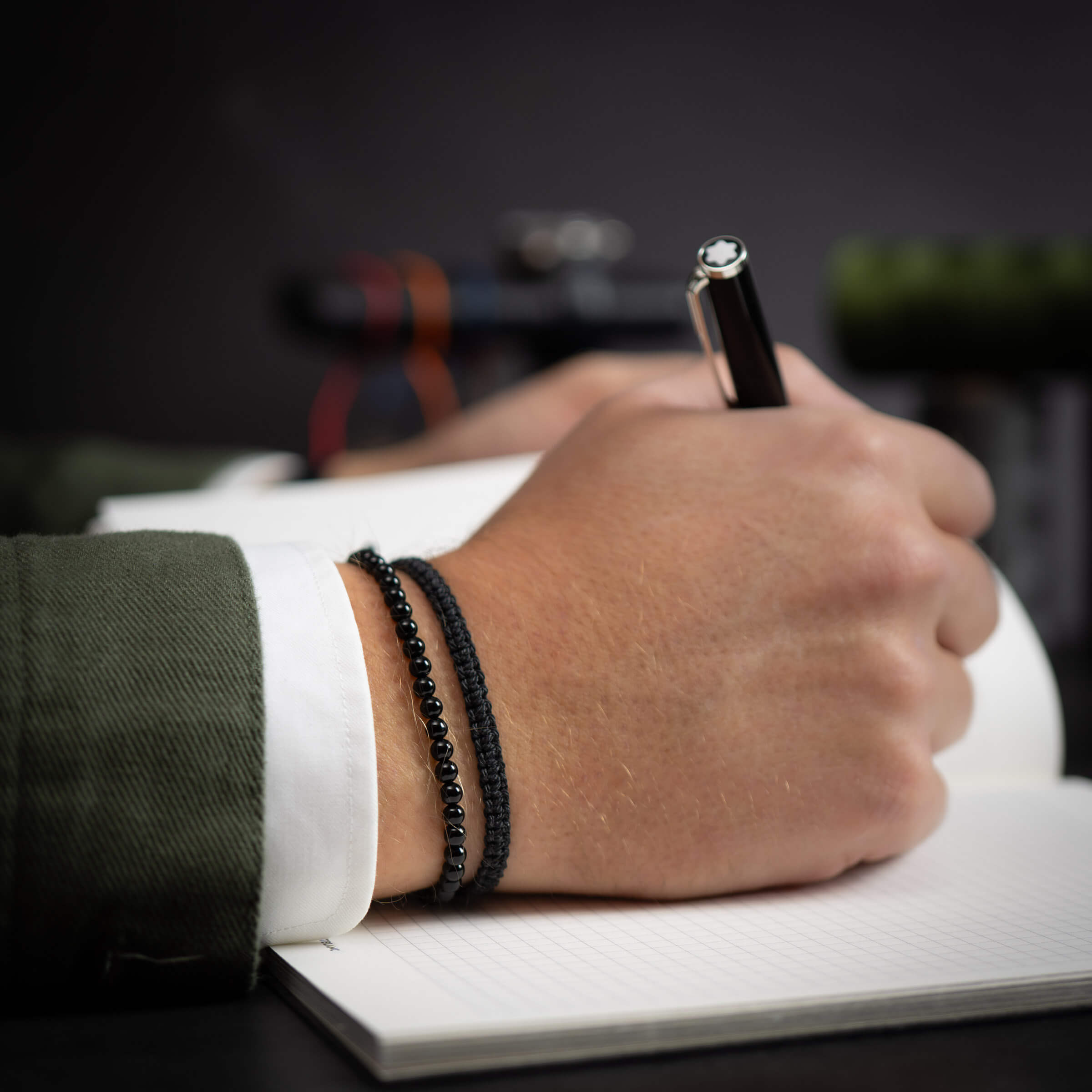 A person's hand wearing a black onyx bracelet and black cobra bracelet is holding a pen, writing on a notepad.