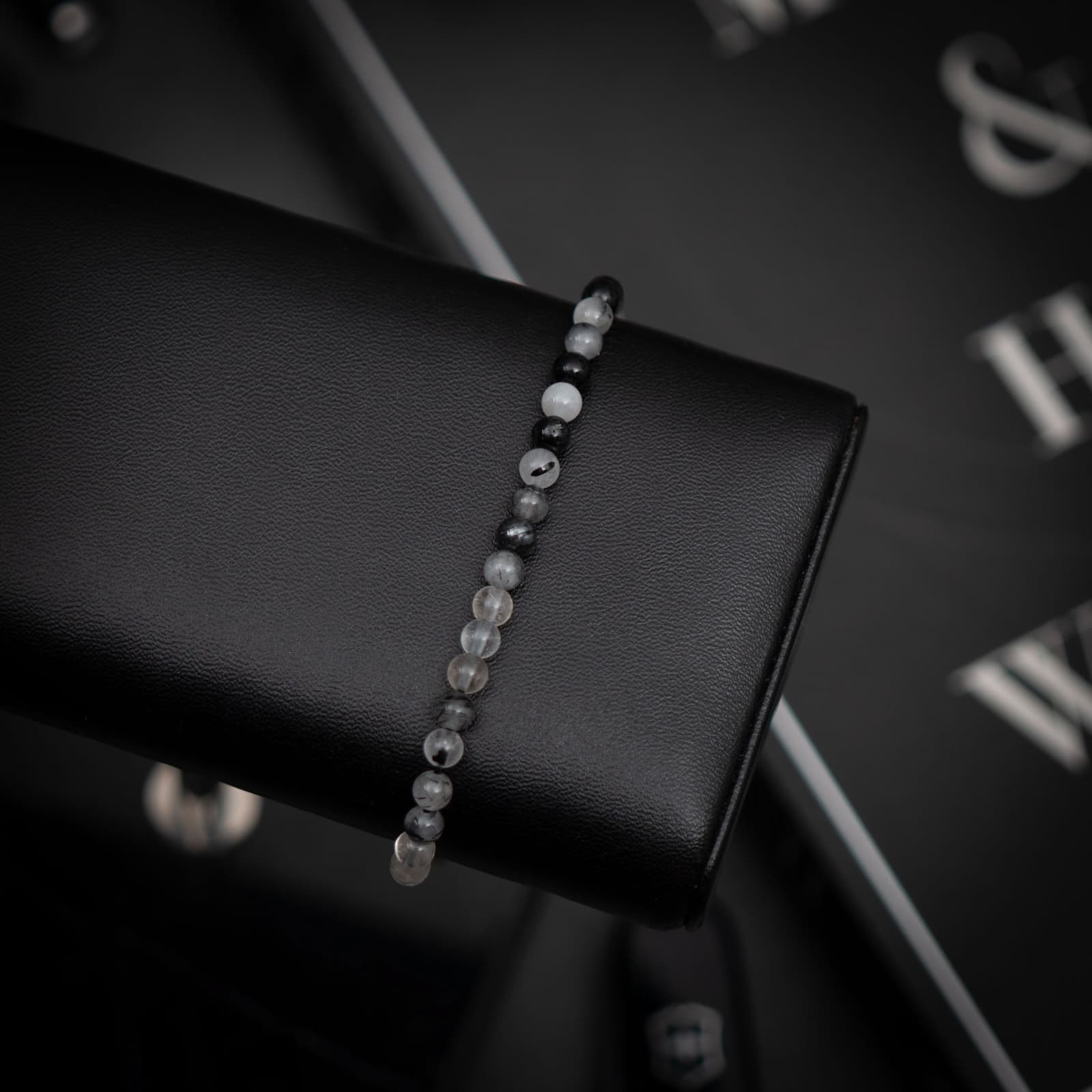 A black rutilated gemstone bracelet displayed on a black stand with a reflection on a glass surface.
