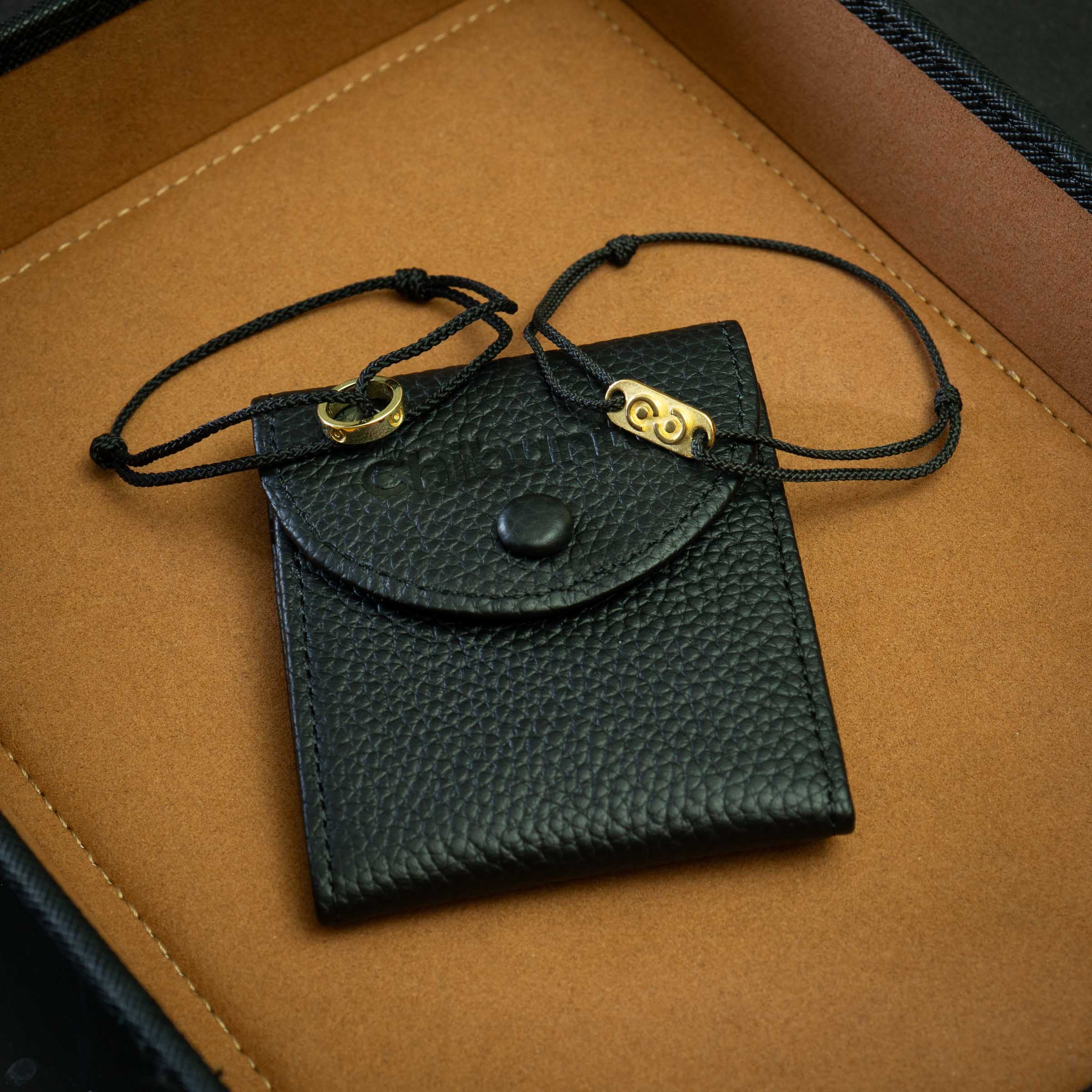 A black 14k gold plated ring bracelet and tag bracelet placed on top of a premium genuine leather bracelet pouch, presented on a wooden surface.