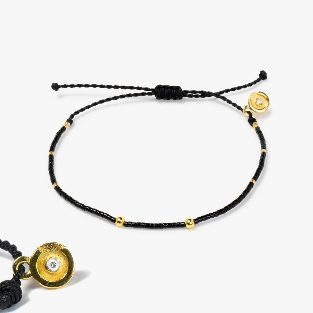 A black bracelet with gold accents and a gold-plated power pose charm with a clear zircon stone in the center, displayed on a white background.