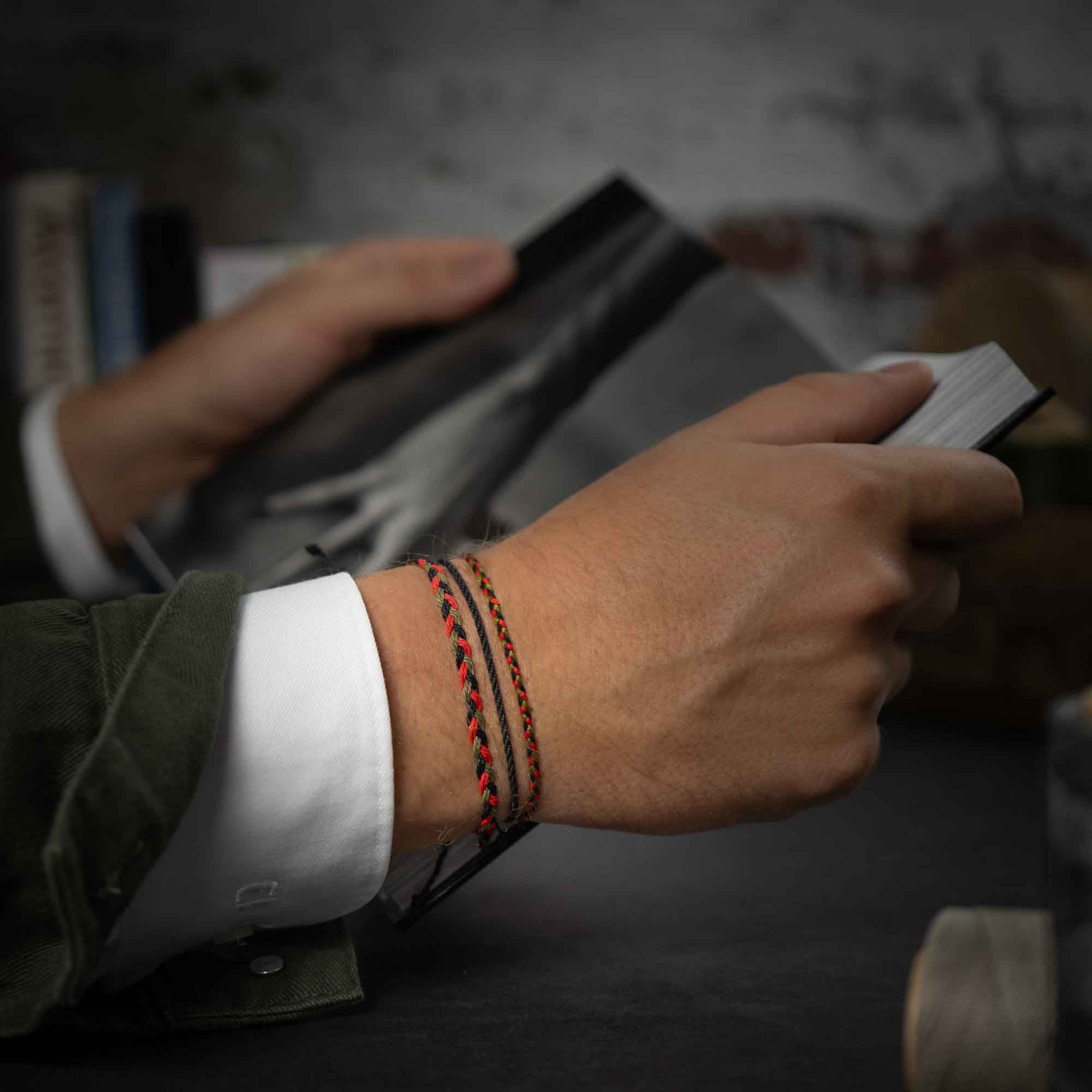 A person's wrist wearing a vintage-style multicolored bracelet, with a gunmetal charm, against a background of books and foliage.