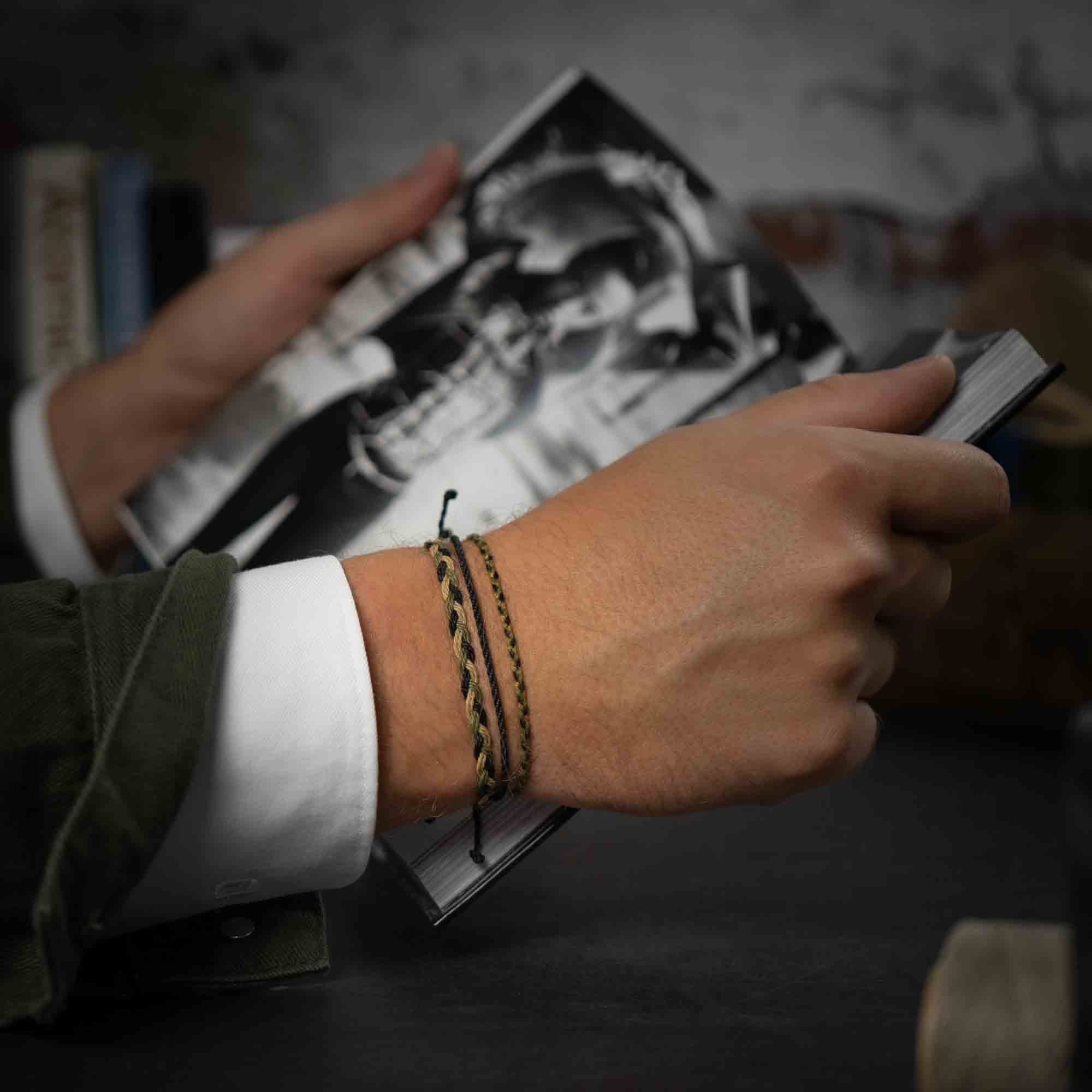 A person's wrist wearing a green and black wax-coated polyester bracelet with a gunmetal charm, while they hold a book.