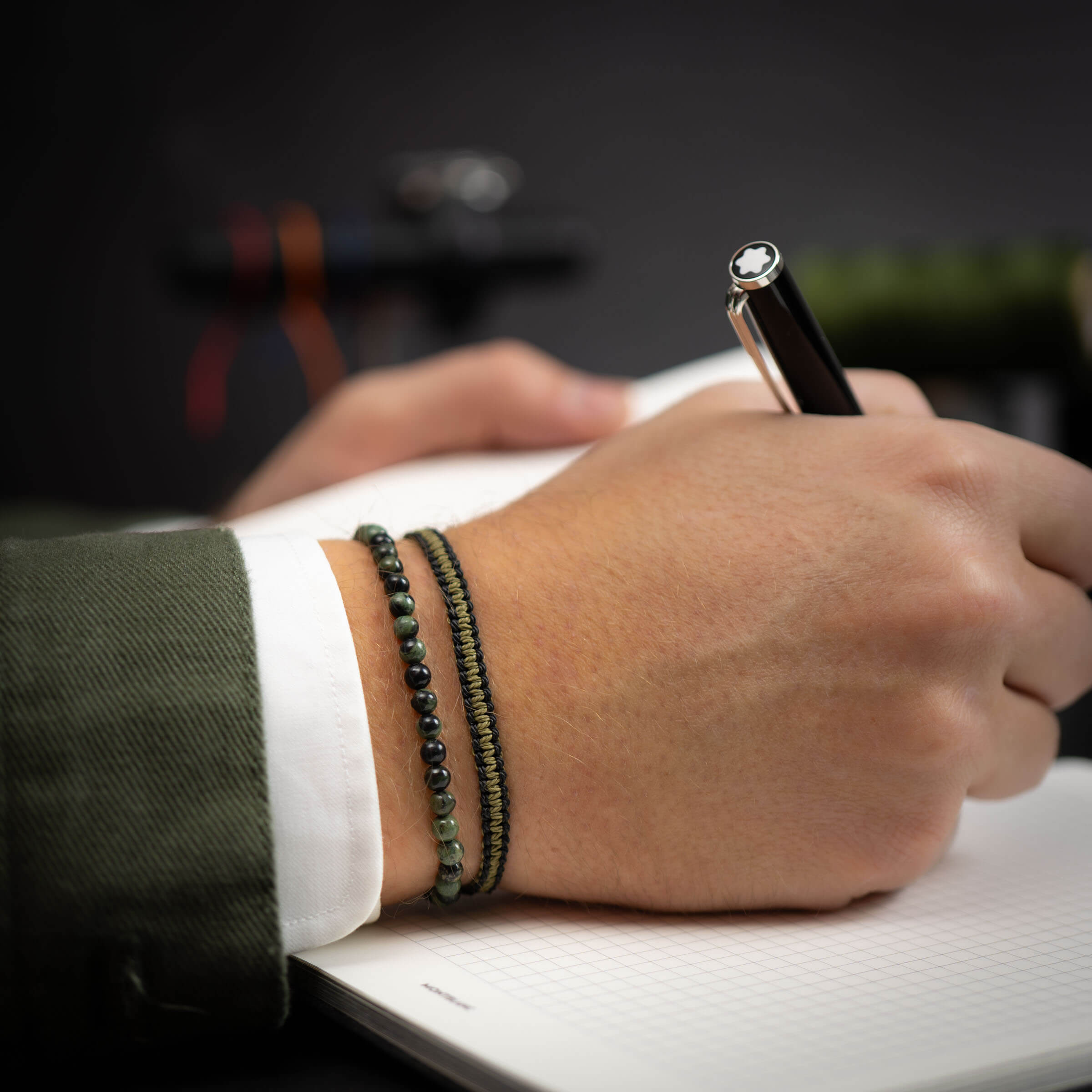 A person's hand wearing a Moss Agate & Military Cobra bracelet with a pen in their hand, writing on a paper.