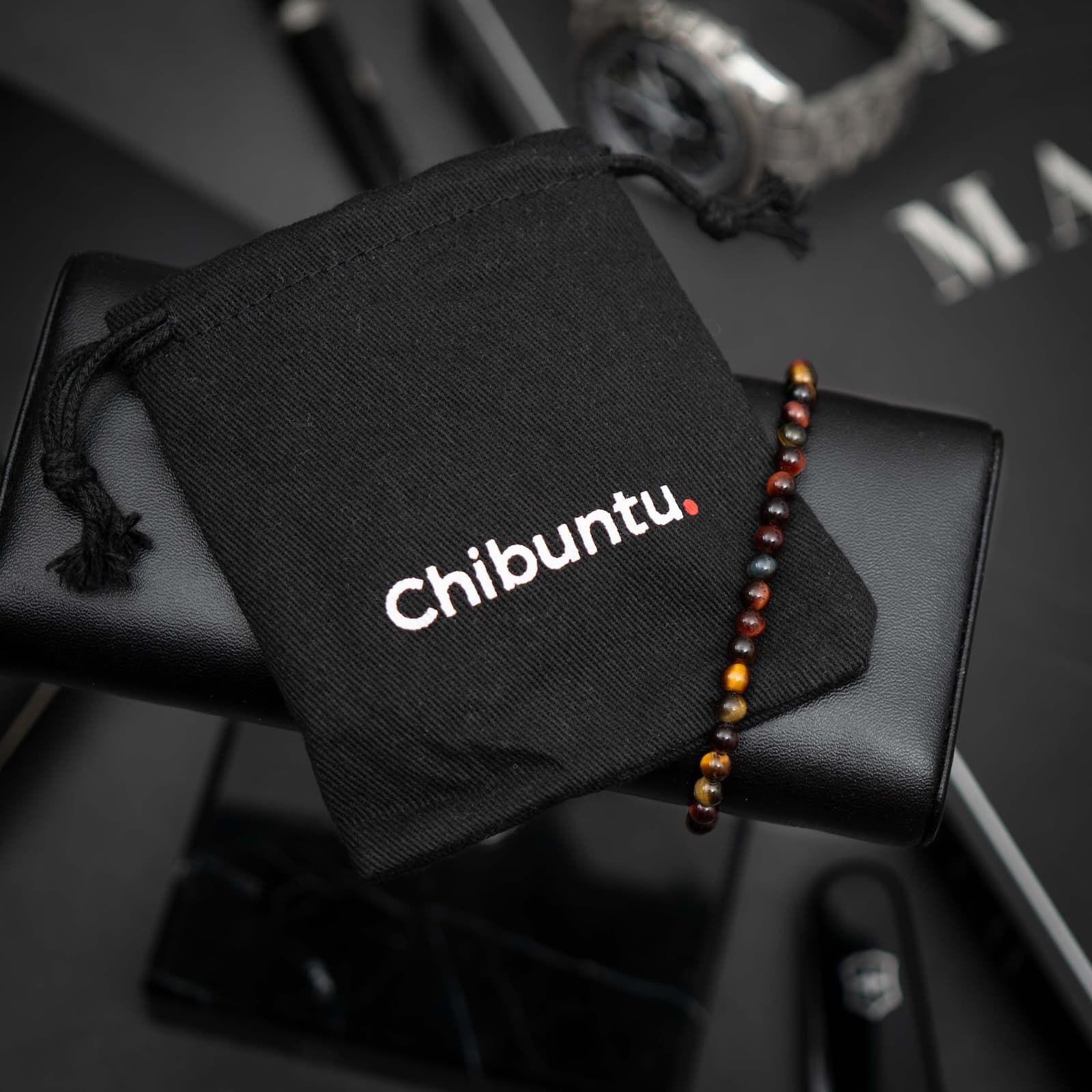 A multicolor tiger gemstone bracelet displayed on a black surface next to a black pouch with the text 'Chibuntu' on it.