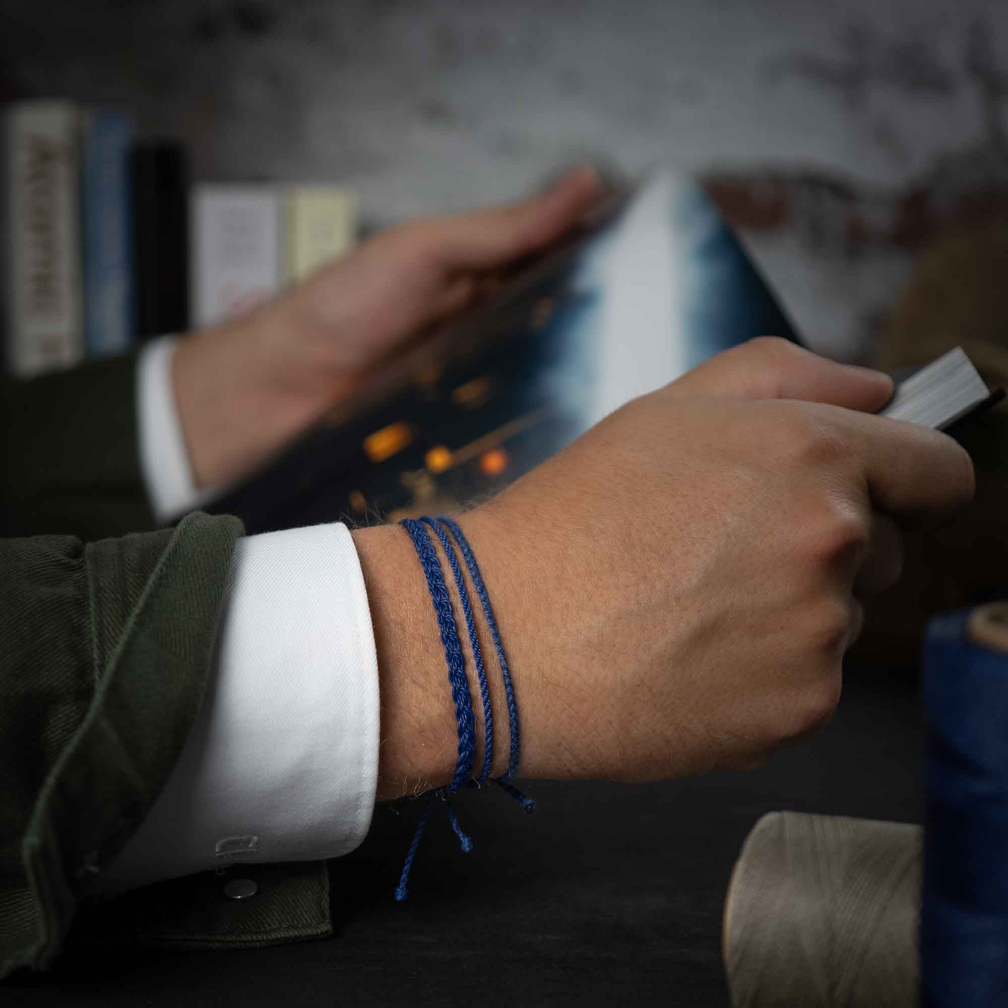A person's wrist wearing a navy blue bracelet with a distinctive pattern, in front of a backdrop suggesting an ocean or beach setting with books and a surfboard.