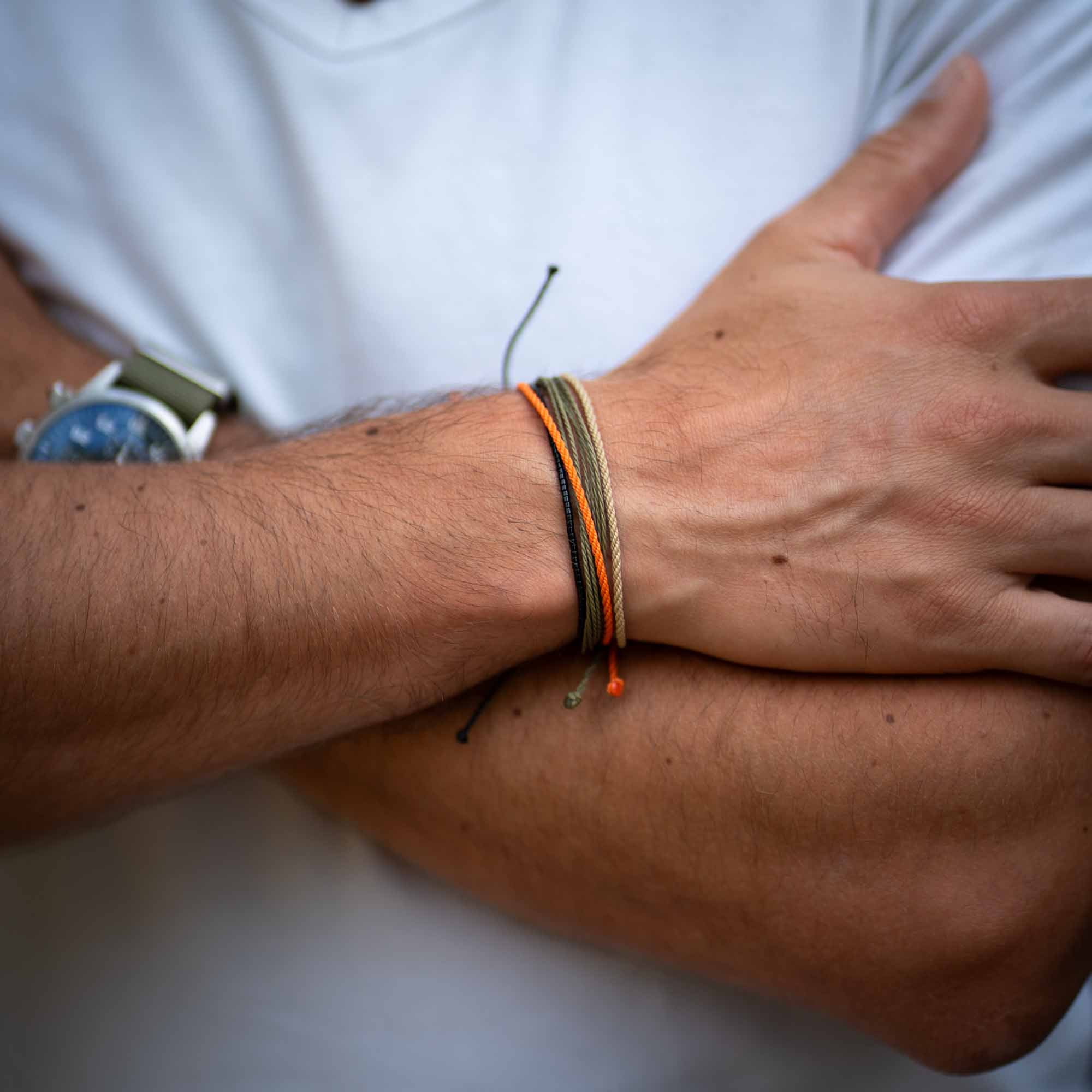 An image of a person wearing three wristbands, two with a wax-coated polyester and one beaded, in earthy colors with vibrant orange, on their forearm.