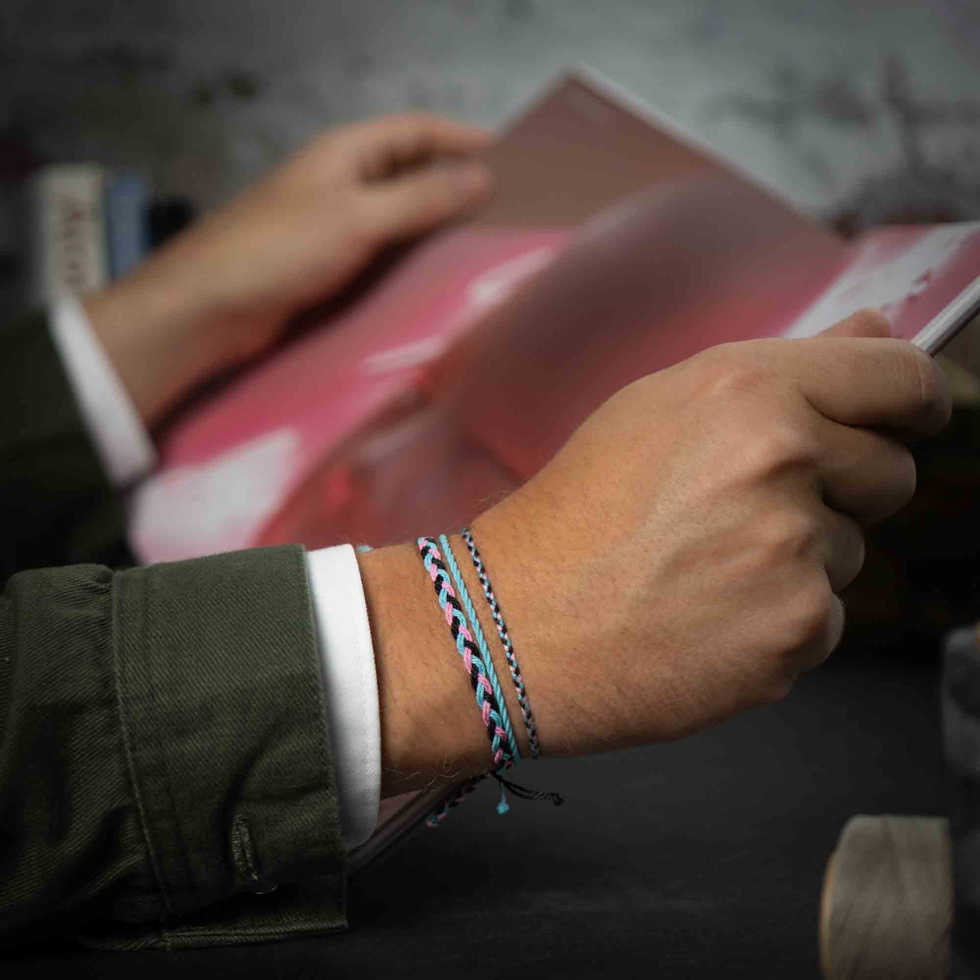 A person's wrist wearing a black and blue wax-coated polyester bracelet with a metallic charm, displayed in front of a red and white object.