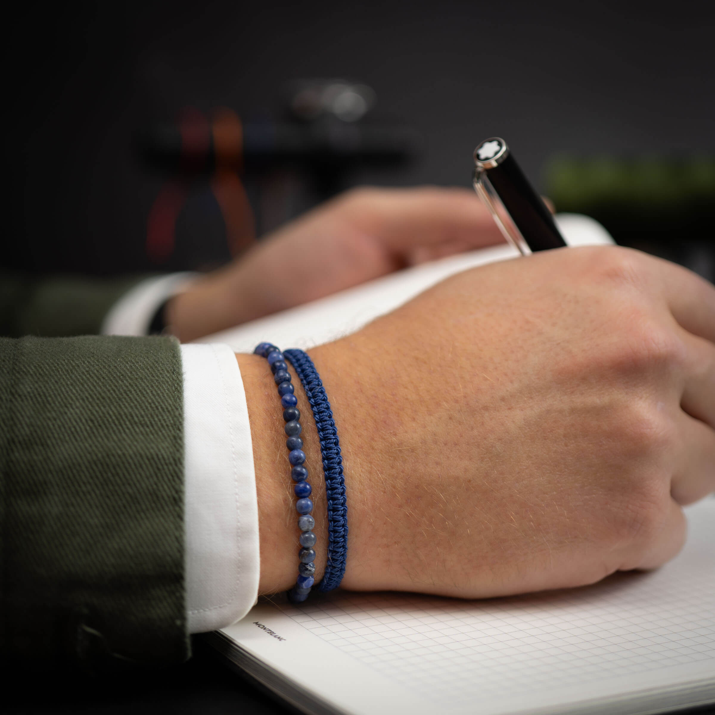 A person's wrist wearing a navy blue cobra bracelet with a Sodalite gemstone bracelet, while writing in a notebook.