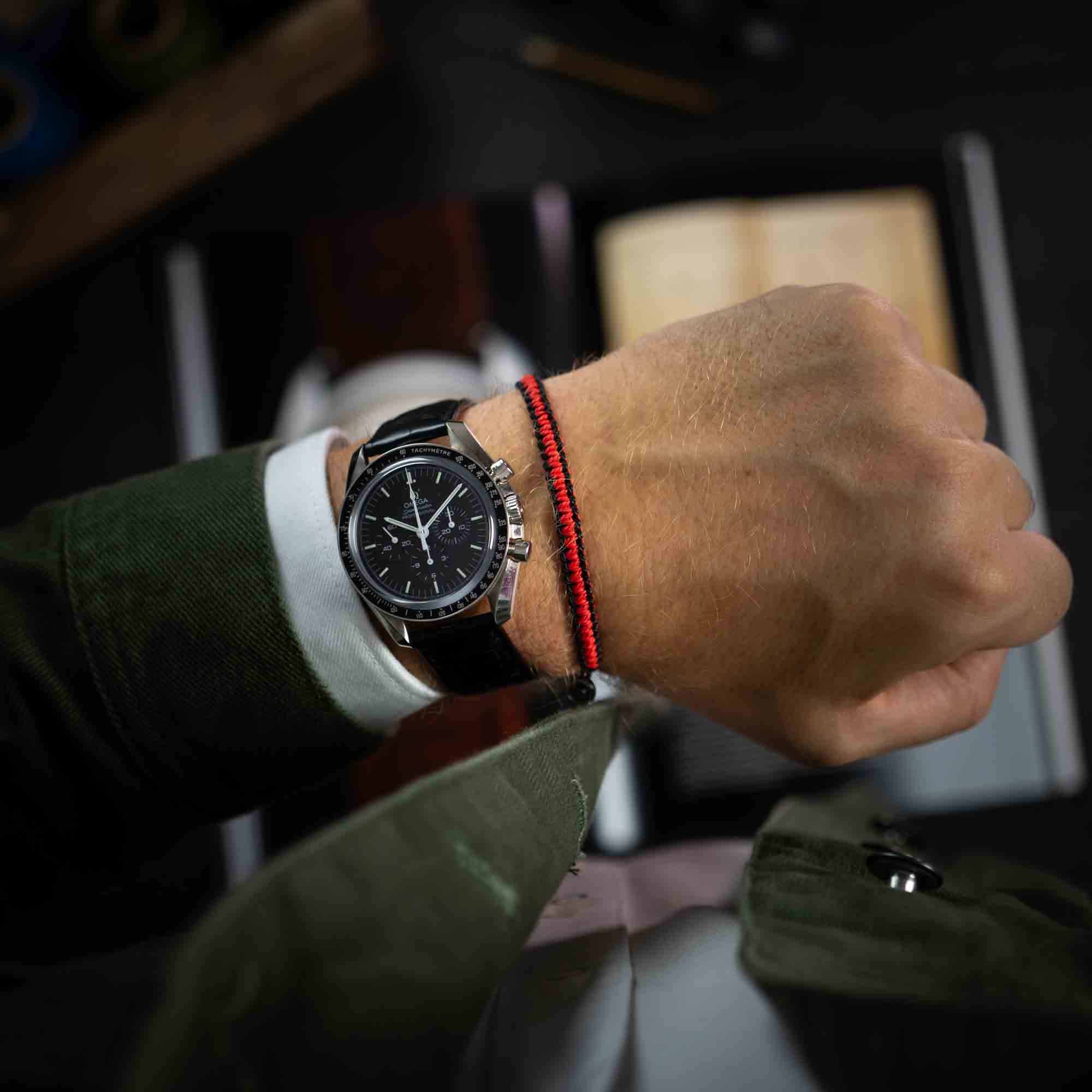 the red and black bracelet for men on the left wrist next to an omega speedmaster