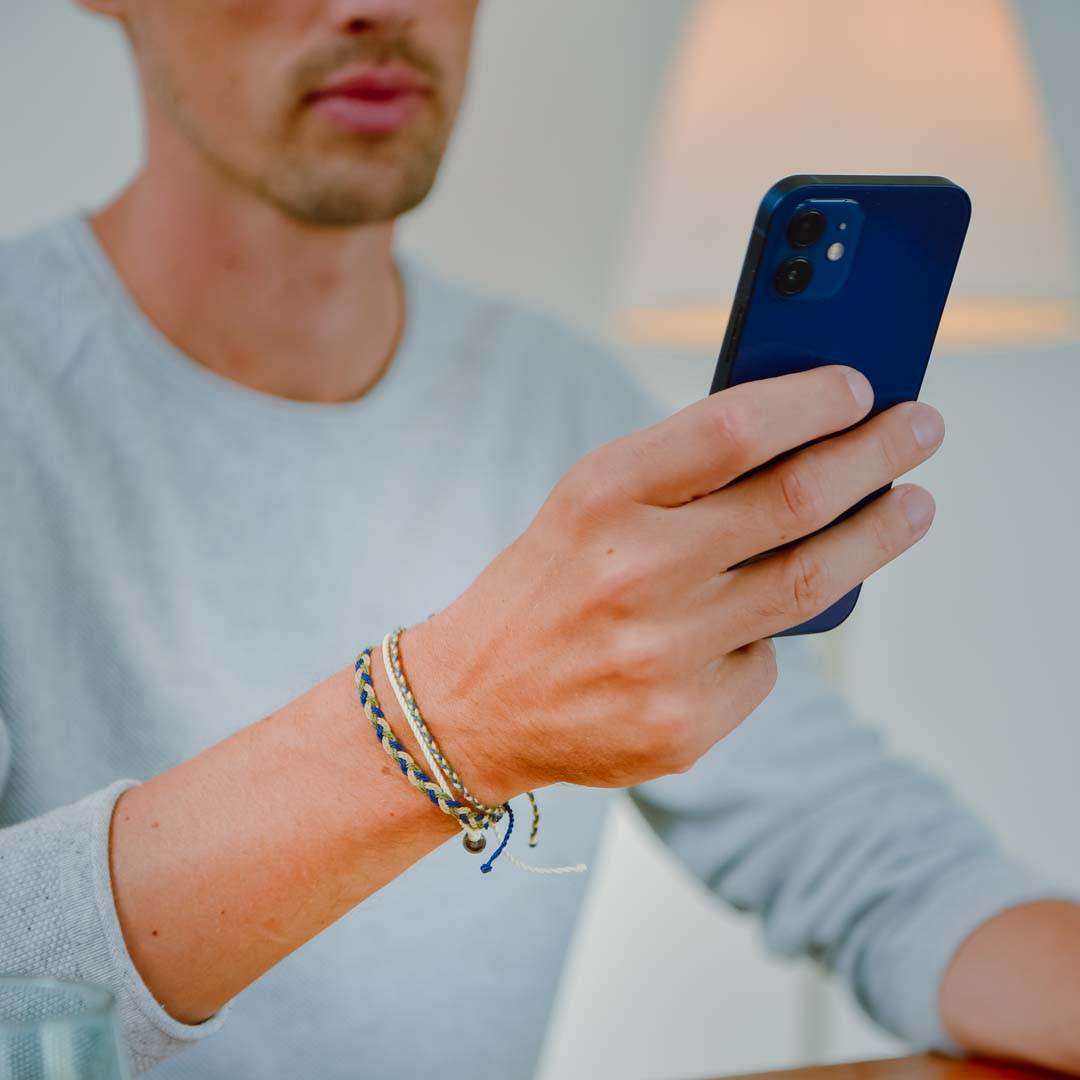 A person is holding a smartphone in their right hand, with a focus on their left wrist wearing a beige and green bracelet pack