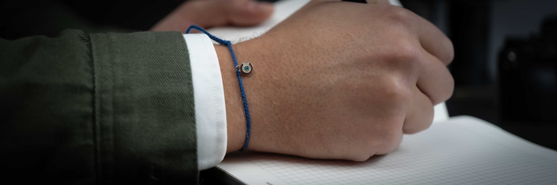Close-up of a person's wrist with a blue bracelet, wearing a green jacket and white shirt.